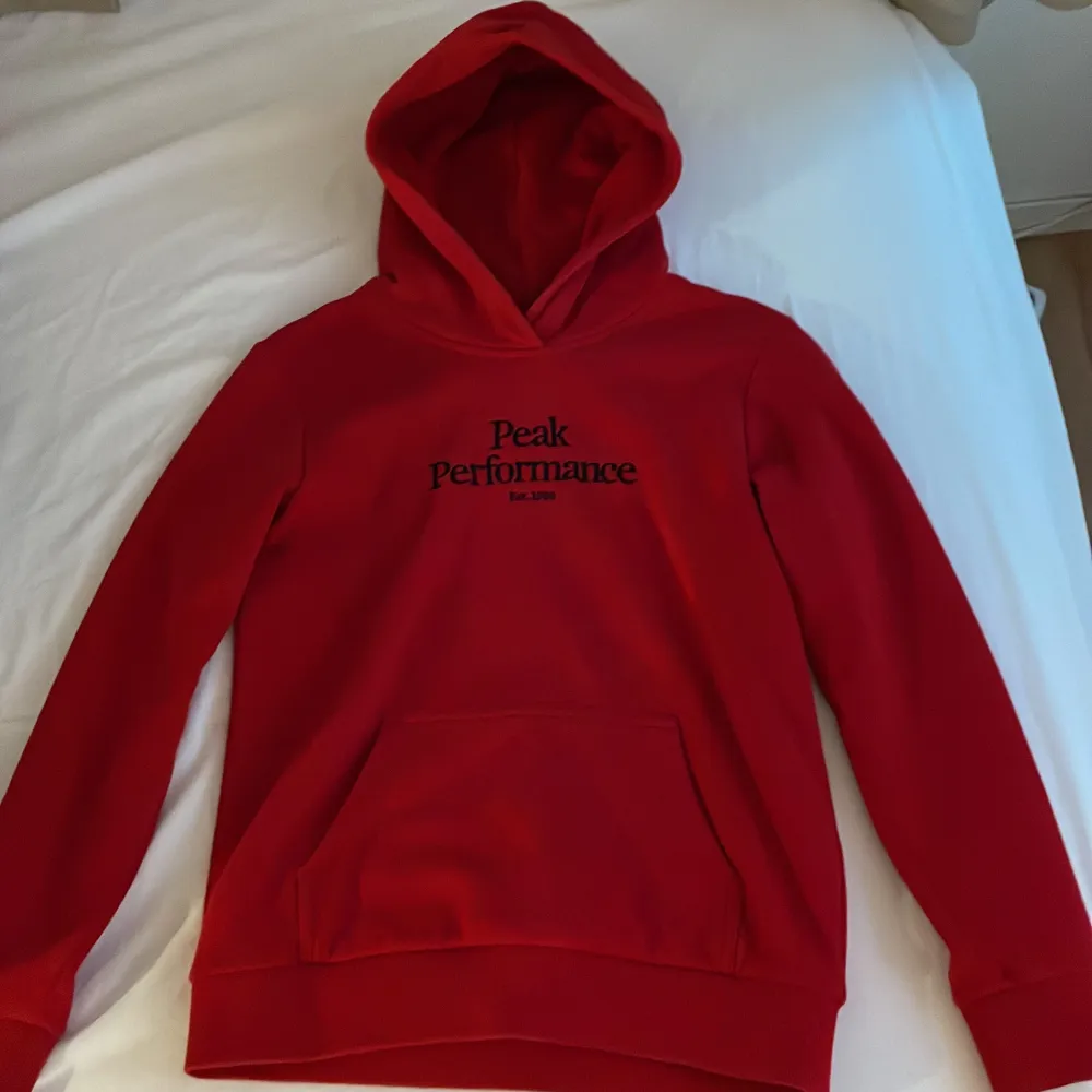 Snygg röd hoodie från Peak Performance med svart logga på bröstet. Den har en klassisk känguruficka och en bekväm huva. Perfekt för en avslappnad stil.. Neuletakit & Villapaidat.