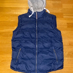 Blå dunväst med grå huva från Cedarwood State - Säljer en blå dunväst från Cedarwood State med en grå huva. Västen har dragkedja framtill och två fickor med dragkedjor. Perfekt för kyligare dagar när du vill hålla stilen.