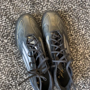 Svarta fotbollsskor från Adidas - Snygga svarta fotbollsskor från Adidas med vita detaljer och snörning. Skorna har en modern design med tydliga ränder och en slitstark sula med dobbar för bra grepp på planen.