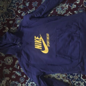Lila hoodie från Nike - Säljer en lila hoodie från Nike med gul logga och texten 'Sportswear' på framsidan. Tröjan har en klassisk känguruficka och justerbar huva med dragsko. Perfekt för en sportig look eller avslappnade dagar.