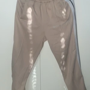 Beige träningsbyxor från Palm Angels - Snygga beige träningsbyxor från Palm Angels med vit och svart rand längs sidan. Byxorna har en elastisk midja och dragkedja vid bensluten. Perfekta för en sportig look.