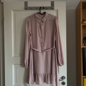 Ljusrosa klänning från NA-KD - Säljer en söt ljusrosa klänning från NA-KD med långa ärmar och knappar framtill. Klänningen har en volangdetalj vid fållen och ett knytband i midjan för en fin passform. Perfekt för vårens och sommarens tillfällen! 🌸