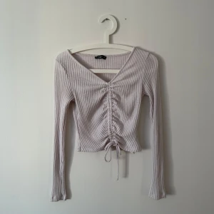 Ljuslila ribbad topp med knyt - Säljer en ljuslila ribbad topp med v-ringning och knyt framtill. Toppen har långa ärmar och en snygg, figurnära passform. Perfekt för en stilren look!