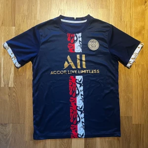 PSG Fotbollströja - Snygg mörkblå fotbollströja med korta ärmar och ett iögonfallande guldtryck på bröstet. Tröjan har ett rött och vitt mönster i mitten och detaljer i guld på ärmsluten. Perfekt för fotbollsfans som vill sticka ut på planen.