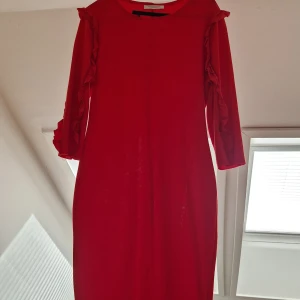 Röd klänning med volangdetaljer - Snygg röd klänning med volangdetaljer längs ärmarna. Klänningen har en enkel och elegant design med en normal passform och trekvartsärmar. Perfekt för att sticka ut med en djärv färg.