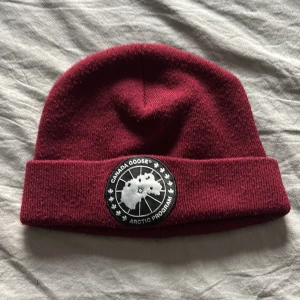 Vinröd mössa från Canada Goose - Säljer en snygg vinröd mössa från Canada Goose med ett stilrent märke framtill. Perfekt för kyliga dagar och ger en trendig look. Mössan är mjuk och bekväm att bära.