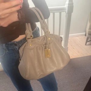 Beige Marc Jacobs väska med gulddetaljer - Så snygg Beige Marc Jacobs väska. Köpte här på Plick för 4000kr för några månader sen. Väskan är i bra skick förutom att en av plupparna på guld plattan har ramlat av så därav lägre pris💕