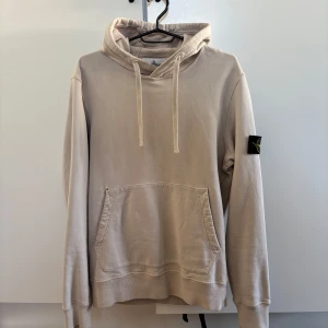 Beige hoodie från Stone Island - Säljer en snygg beige hoodie från Stone Island med klassisk logga på ärmen. Den har en känguruficka och justerbar huva med snören. Perfekt för en stilren och avslappnad look. Obs. Litet hål vid fickan.