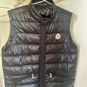 Svart dunväst från Moncler - Snygg svart dunväst från Moncler med dragkedja och hög krage. Västen har två praktiska fickor med dragkedja och Monclers logga på bröstet. Perfekt för vår, sommar och höst. Utbytt dragkedja hos skräddare. Storlek 14 år (164)