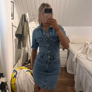 Jeansklänning med knappar - Snygg jeansklänning med knappar framtill och korta ärmar. Klänningen har en klassisk krage och två bröstfickor. Perfekt för en avslappnad stil med en touch av denim.