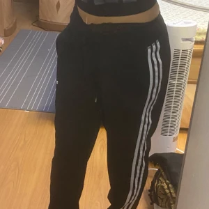 Svarta träningsbyxor från Adidas - Snygga svarta träningsbyxor från Adidas med de klassiska tre vita ränderna längs sidorna. Byxorna har en bekväm passform och är perfekta för träning eller en avslappnad stil. Adidas-loggan är diskret placerad på ena benet.