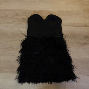 Svart axelbandslös klänning med fjädrar - Snygg svart axelbandslös klänning med fjäderdetaljer. Perfekt för en kväll ute eller en speciell tillställning. Klänningen har en tight passform som framhäver figuren.