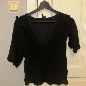 Svart omlottopp med volangdetaljer - Snygg svart omlottopp med volangdetaljer och trekvartsärm. Perfekt för en stilren look. Toppen har en v-ringad design som ger en elegant touch.