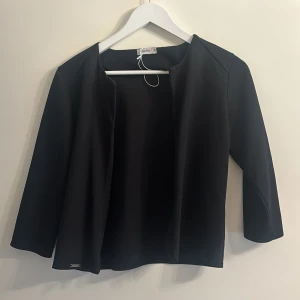 Svart kavaj från Gerry Weber - Elegant svart kavaj från Gerry Weber med en stilren design. Kavajen har långa ärmar och en öppen front utan knappar eller dragkedja. Perfekt för en chic och professionell look.