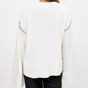 Vit stickad tröja med svarta detaljer - Säljer en stilren vit stickad tröja med svarta randiga detaljer vid axlarna. Tröjan har en avslappnad passform med långa ärmar och breda ribbade muddar vid ärmslut och nederkant. Helt oanvänt med lappen kvar