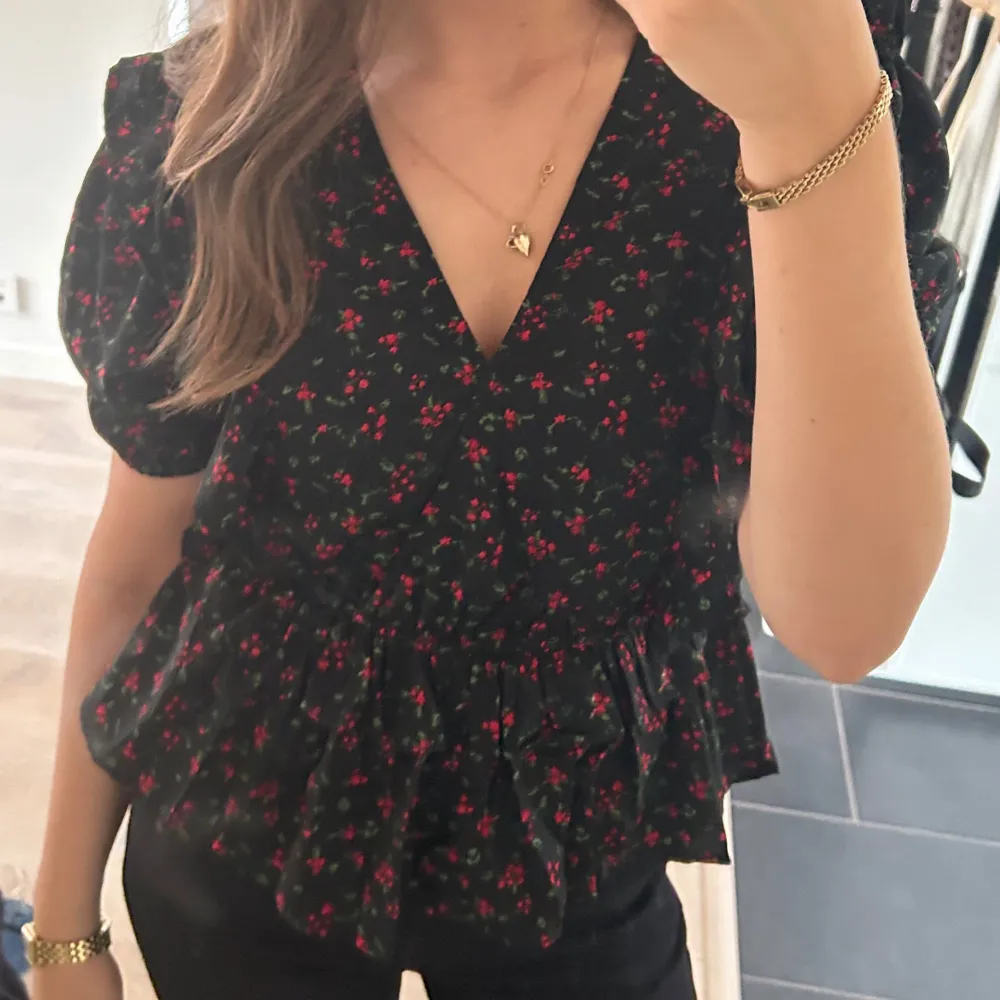 Säljer en svart blus med små röda blommor och gröna blad. Blusen har korta puffärmar och en v-ringad design med volangdetaljer i nederkant. . Yläosat.