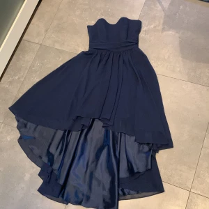 Mörkblå axelbandslös klänning från Swing - Säljer en elegant mörkblå axelbandslös klänning från Swing. Klänningen har en vacker asymmetrisk design med längre bakdel och är perfekt för speciella tillfällen. Den har en dragkedja i ryggen för enkel påklädning.