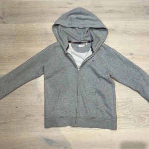 Grå hoodie från GANT - Säljer en stilren grå hoodie från GANT med dragkedja och två fickor framtill. Den har en klassisk design med ribbade muddar och en liten logga på bröstet. Perfekt för en avslappnad look.