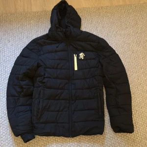Svart quiltad jacka med huva Gymking - Säljer en svart quiltad jacka med huva och dragkedja. Jackan har en vit dragkedja på bröstet och ett broderat märke. Perfekt för kyliga dagar.