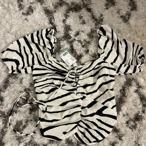 Zebramönstrad topp med puffärmar - Snygg zebramönstrad topp med puffiga kortärmar och snörning framtill. Perfekt för en trendig look med det svartvita mönstret och unika designen. Passar bra till både jeans och kjol.