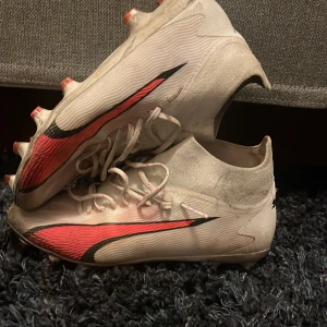 Fotbollsskor med snörning - Säljer ett par fotbollsskor i vitt och rött med snörning. Skorna har en strömlinjeformad design med röda och svarta detaljer på sidorna. Perfekta för fotbollsplanen med sina dobbar för bra grepp.