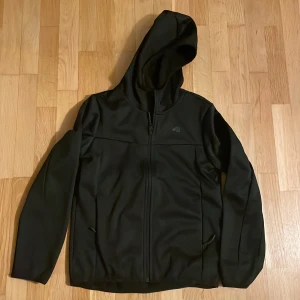 Svart jacka med huva från 4F - Säljer en svart vindjacka med huva från 4F. Jackan har en dragkedja framtill och är långärmad. Perfekt för kyligare dagar med sitt mjuka foder.