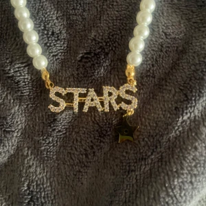 Halsband med pärlor och stjärndetalj - Elegant halsband från en liten butik i Belgien med vita pärlor och en gnistrande 'STARS'-text i guld. Halsbandet har en stjärnberlock som hänger nedanför texten. Perfekt för att ge en glittrig touch till din outfit. Priset kan diskuteras 