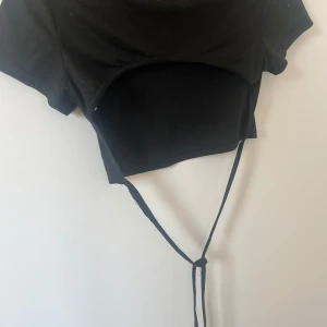 Svart croppad topp med knytband - Snygg svart croppad topp med korta ärmar, knytband i midjan och öppen rygg. Perfekt för en trendig look. 