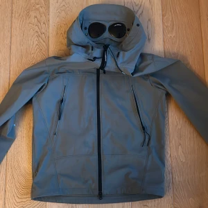kom med byten! - Säljer en grön vindjacka från C.P. Company med ikoniska glasögon i huvan. Jackan är tillverkad i mjukt skaltyg med stretch och återvunnen nylon. Den har dragkedja framtill och flera praktiska fickor.