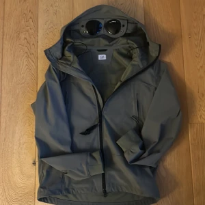 Grön vindjacka från C.P. Company - Säljer en grön vindjacka från C.P. Company med huva och värsting glasögon. Jackan har dragkedja framtill och är tillverkad i ett stretchigt soft shell-material av återvunnen nylon. Perfekt för en stilren look.