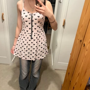 Rosa linneklänning med rosett-mönster - Söt rosa linneklänning med svarta rosetter och dragkedja framtill. Perfekt för en avslappnad dag eller en somrig utflykt. Tunna axelband och en luftig passform gör den bekväm att bära.