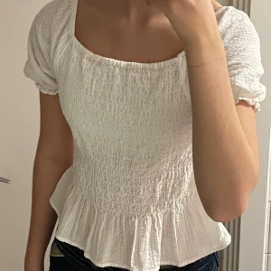 Vit blus från Kappahl - Säljer en söt vit blus från Kappahl med korta puffärmar och volangdetalj i nederkant. Är i nytt skick, utan defekter! Toppen har en smockad struktur som ger en fin passform. Perfekt för sommarens varma dagar!