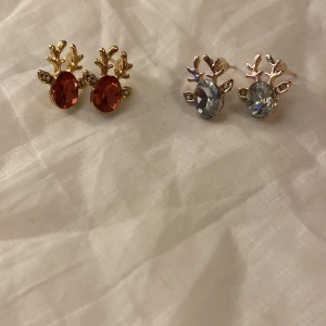 Örhängen med renhornsdesign - Säljer två par örhängen med renhornsdesign. Ett par har röda stenar och guldfärgade detaljer, medan det andra paret har klara stenar med silverfärgade detaljer. Perfekta för att ge en festlig touch till din outfit!