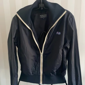 Svart bomberjacka från Polo Jeans Company - Snygg svart bomberjacka från Polo Jeans Company med dragkedja och broderad logga på bröstet. Jackan har långa ärmar och ribbade muddar vid ärmslut och nederkant. Perfekt för en stilren look.