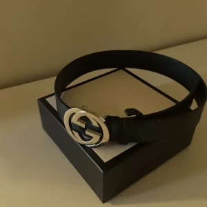Svart bälte med silverfärgat spänne - Snyggt svart bälte från Gucci med ett iögonfallande silverfärgat spänne i form av märkets ikoniska logotyp. Perfekt för att ge en stilren touch till din outfit. Bältet är tillverkat i skinn och har en klassisk design som passar till många olika stilar.