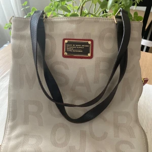 Beige axelväska från Marc by Marc Jacobs - Snygg beige axelväska från Marc by Marc Jacobs med tryckt logotypmönster. Väskan har svarta axelremmar och en praktisk dragkedja på baksidan. Inuti finns en röd etikett med märkesnamnet. Perfekt för att bära dina dagliga nödvändigheter med stil.