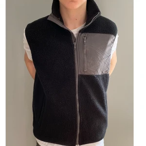 Svart fleeceväst med grå detaljer - Snygg svart fleeceväst med grå detaljer och dragkedja framtill. Perfekt för lager-på-lager under kyliga dagar. Västen har en praktisk bröstficka med dragkedja för säker förvaring. Storlek: S 