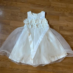 Vit ärmlös klänning med blommor - Säljer en söt vit klänning med stora blommor och pärldetaljer runt halsen. Klänningen är ärmlös och har en vacker volangkjol som ger en fluffig look. Perfekt för speciella tillfällen! 💐 Super söt och är i mycket bra skick. 