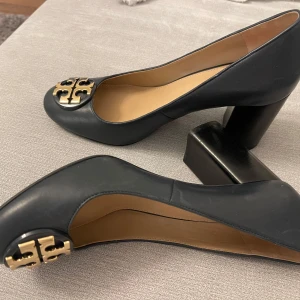 Svarta Tory Burch skor med gulddetalj - Eleganta svarta pumps från Tory Burch i skinn med en stilren gulddetalj framtill. Skorna har en bekväm klack och rundad tå, vilket gör dem perfekta för en chic look.  Skorna är äkta , knappt använda och är i storlek 8. 