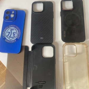 Samling av mobilskal för iPhone 14 pro max - Säljer en samling av olika mobilskal för iPhone. Inkluderar ett blått skal med Leksands IF-logga, ett kolfibermönstrat skal, ett transparent skal och flera svarta skal. Perfekt för att byta stil på din telefon när du vill!