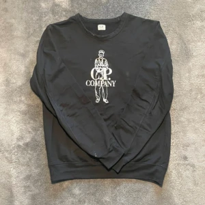 Svart tröja från C.P. Company - Säljer en svart tröja från C.P. Company med ett coolt tryck av en figur och texten 'C.P. Company' på framsidan. Tröjan har långa ärmar och en rund halsringning. Perfekt för en avslappnad stil.