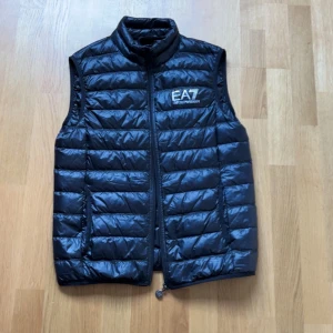 Svart dunväst från Emporio Armani - Snygg svart dunväst från Emporio Armani med dragkedja och hög krage. Perfekt för kyligare dagar och ger en stilren look. Västen har ett EA7-märke på bröstet och quiltad design.