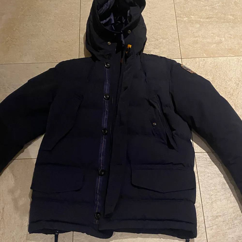 Säljer en snygg mörkblå dunjacka från Parajumpers. Jackan har en huva och stängs med både dragkedja och knappar. Den har flera praktiska fickor och en cool patch på ärmen. Perfekt för kalla dagar! 🧥. Takit.