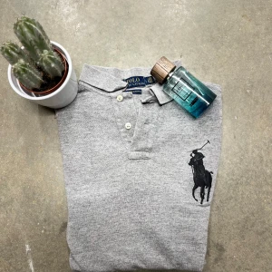 Grå pikétröja från Ralph Lauren - Säljer en stilren grå pikétröja från Ralph Lauren med klassisk krage och knappar framtill. Tröjan har en broderad logga på bröstet och en siffra på ärmen. Perfekt för en avslappnad men ändå snygg look.