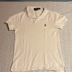 Beige pikétröja från Ralph Lauren - Säljer en stilren beige pikétröja från Ralph Lauren i slim fit. Tröjan har korta ärmar och en klassisk krage med knappar. Den är dekorerad med det ikoniska broderade logotypen i mörkblått på bröstet. Perfekt för en avslappnad men ändå elegant look.