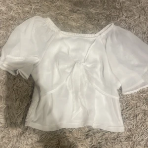 Vit blus från Lindex - Säljer en söt vit blus från Lindex med puffiga korta ärmar och en knytning framtill. Blusen har en elastisk detalj vid midjan för en bekväm 