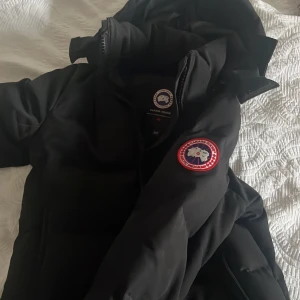 Svart dunjacka från Canada Goose - Säljer en svart dunjacka från Canada Goose. Jackan har en klassisk design med huva och dragkedja framtill. Perfekt för kalla vinterdagar. Den har två stora fickor framtill och en broderad logotyp på ärmen.