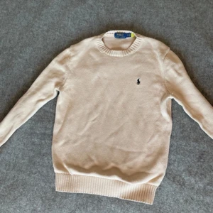 Beige tröja från Polo Ralph Lauren - Säljer en stilren beige tröja från Polo Ralph Lauren med rund hals och det klassiska logotypbroderiet på bröstet. Tröjan har långa ärmar och ribbade muddar vid ärmslut och nederkant. Perfekt för en avslappnad men snygg look.