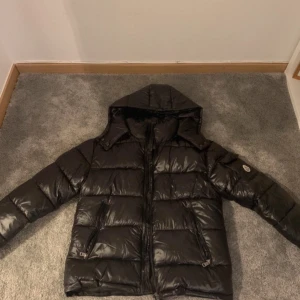 Svart dunjacka med huva - Säljer en svart dunjacka med huva. Jackan har en glansig yta och är långärmad med dragkedja framtill. Perfekt för kyliga dagar.