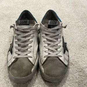 GGDB sneakers med stjärndetalj - Snygga GGDB sneakers i vitt med en svart stjärna på sidan. Skorna har en klassisk snörning och en cool, sliten look. Den blå detaljen vid hälen ger en extra touch. Perfekta för en avslappnad stil.
